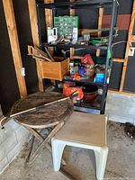 Full view of the metal shelving unit with several items including hand tools, containers, and gardening supplies, along with a white plastic patio chair and small round folding wooden table in front.