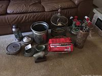 Photo showing the entire lot on floor in front of couch, including decorative bird cage, planter with flowers, brass fireplace pot, pewter bison figure, fireplace logs, and various metal lanterns and containers.