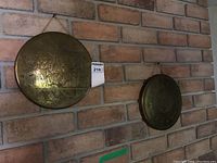 Two round copper wall plates hanging on a brick wall, showing etched designs and hanging loops.