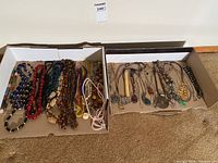Two cardboard boxes containing a variety of costume jewelry including bead necklaces of various colors and styles in the left box, and chain necklaces with different pendants in the right box.