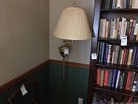 Full view of brass-look floor lamp positioned near bookshelf, showing pleated beige lampshade and extendable arm.