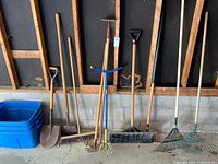 Full view of garden tools lined up against a wall with three blue rubber containers to the left.