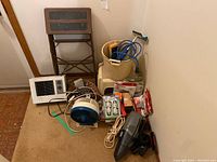 Photo showing small step ladder, small plastic step stool, Dirt Devil vacuum cleaner, small fan, replacement vacuum bags, and various light bulbs and tools on a carpeted floor against a beige wall.