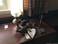 Overview of lot items on wooden table including oil lamp, carved whale, brass bell, totem pole figure, frames, and heart wall hanging.