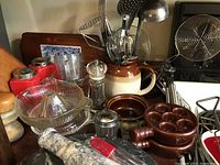 Photo showing an assortment of kitchen items including glass juicers, escargot server, salt and pepper shakers, ceramic utensil crock, and wooden and metal utensils.