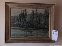 Framed oil painting showing a natural outdoor scene with trees, water, and distant hills.