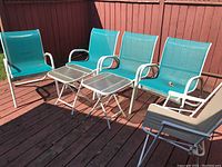 Four teal blue metal frame stacking chairs, two small folding tempered glass top tables, one white folding chair on a wooden deck.