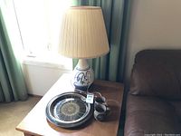 Full view of the pottery table lamp on a wooden side table with pleated fabric shade, large round platter on table, and pair of pottery mugs beside the lamp.
