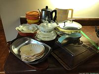Full view of retro kitchen collection including Tupperware, serving bowls, metal coffee percolator, casserole dishes, shell shaped plates, and serving trays on counter.