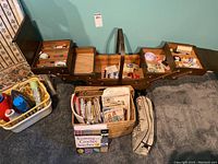 Photo showing wooden sewing box fully opened with compartments of threads, buttons, needles, and other sewing notions along with wicker basket and knitting books and supplies on floor.
