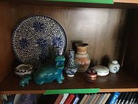Ceramic and pottery collection displayed on a wooden shelf including blue and white platter, turquoise hippos, pottery vase, ginger jar, pierced vases, and ashtray
