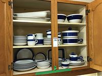 Kitchen cabinet with multiple shelves holding the dinnerware set in cream with blue ring pattern, showing plates, bowls, cups, and serving dishes.