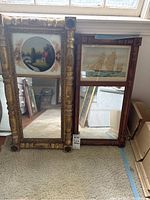 Two antique framed mirrors side by side by a window. Left mirror features a painted house scene and gold ornate frame. Right mirror has a sailing ship image and dark wooden frame.
