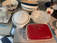 Photo showing a variety of covered casseroles and pie plates, including a red ceramic casserole, clear glass casserole, and a white casserole with decorative trim, along with white fluted pie plates.