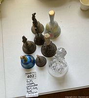 Seven assorted bells arranged on white surface showing various materials including metal, porcelain, and glass. Includes visible porcelain bell with blue glaze and figurine handle.