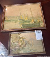 Both framed oil paintings of harbor and boat scenes displayed together on table
