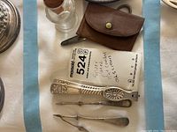 Photo showing silver plated nutcracker, brown leather pouch, old bottle with cork stopper, and several silver plated tools including scissors and picks.