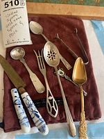 Full lot of vintage flatware laid out on cloth showing spoons, fork, and knife with porcelain handles, some slotted spoons including a gold spoon.