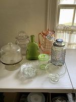 Photo showing all items on table: covered glass jars, green depression glass violin bottle, pink ice tea pitcher, scallop dishes, small glass bowl.