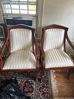 Pair of dark red wood armchairs with attached striped white cushions on seat and back, placed on a patterned rug in front of a window.