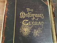 Close-up of the hardcover front cover of The Masterpieces of German Art book showing black and brown leather with gold embossed lettering and decoration, showing wear and weathering.