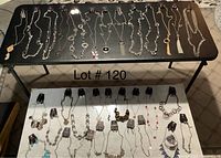 Wide shot of two tables showing 32 necklaces laid out horizontally, necklaces mostly silver-tone with some blue, pearl and pendant features.