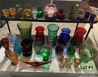 Full lot of colored glassware including vases, bottles, glasses, and decorative figurines arranged in rows on tabletop.