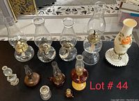 Front view of all 11 lamps arranged on a black surface, showing 5 clear glass oil lamps with chimneys, 2 small oil lamps missing tops, and a decorative milk glass lamp with orange flower design.