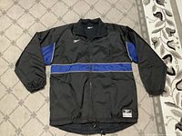 Exterior of black Reebok Legacy coat/jacket with blue panels and white piping on a carpeted floor.