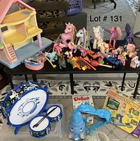 Photo showing small dollhouse, multiple My Little Ponies, toy wind instruments, and toy cars arranged on a table with drums and boxed toys below.