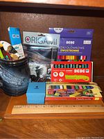 Front view showing the ceramic mug filled with pens, origami paper, pastel boxes, colored pencils, and wooden ruler on the shelf.