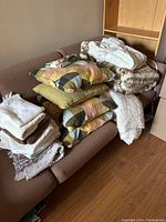 Overall lot displayed on brown sofa including pillows, blankets, throws, and towels stacked