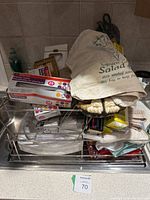 Metal dish drainer rack holding multiple kitchen textiles including pot holders and wiping cloths, rolls of plastic wrap, and a fabric bag with printed text.