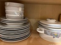White ceramic dishware set including stacked dinner plates, smaller plates, bowls, and a lidded casserole dish with floral design.