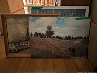 Three framed art reproductions including a rural field scene with flowers and sky, a winter church scene, all framed and leaning on floor.