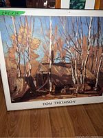Large framed artwork with birch trees labeled 'Tom Thomson' and size tag on upper left corner, resting on wooden floor.
