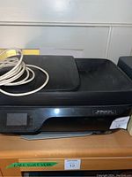 Front and top view of the black HP OfficeJet 3830 printer with coiled power cable on top and LCD control panel visible.