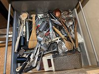 Photo showing a drawer filled with assorted kitchen utensils including graters, wooden spoons, metal cutlery, and peelers mixed together.