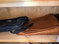 Brown and black leather computer bags stacked on a wooden shelf, brown bag on bottom and black bag on top showing front and straps.