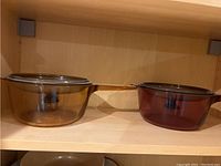 Two Pyrex glass pots with handles on a wooden shelf, one amber and one dark brown color