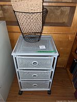Front view of the white metal rolling storage unit with three mesh drawers and black caster wheels. Wire basket visible on top.