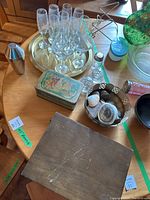 Wide view of lot items including glassware set, cocktail shaker, silver tray, salt and pepper shakers, tin box, and basket with shells
