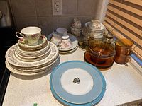 Wide view of vintage porcelain and china collection placed on countertop. Various cups, saucers, plates and amber glass items visible.