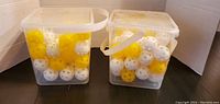 Two transparent square plastic containers with lids filled with white and yellow perforated plastic practice golf balls against a white background.