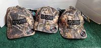 Three camo baseball hats with elone logo on front, new with tags, displayed side by side on green carpet