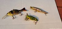 Photo showing three used fishing lures on a white surface, including a segmented fish-shaped lure, a green frog-style lure, and a small minnow-style lure.