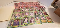 Photo showing 46 baseball cards laid out in rows, mostly in plastic sleeves featuring various baseball players mostly in green bordered cards with some red bordered cards in background.