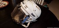 Side view of white Cooper hockey helmet with clear visor, ventilation holes, and chin strap.