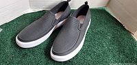 Pair of dark grey slip-on summer shoes with perforations on the sides and top, sitting on green carpet inside a box.