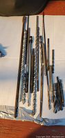 Photo showing a set of large used drill bits of various lengths and diameters placed on a white surface.